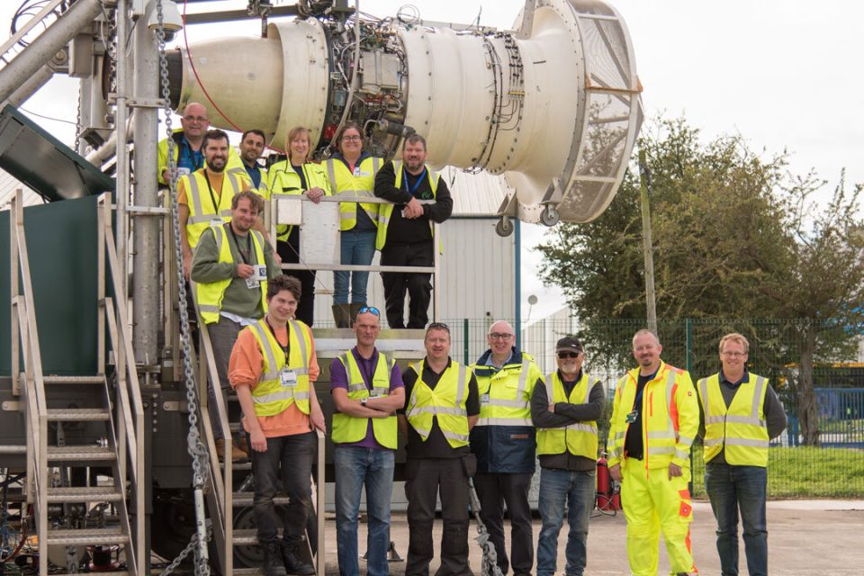 Group of people wearing high visibility jackets stand next to jet engine